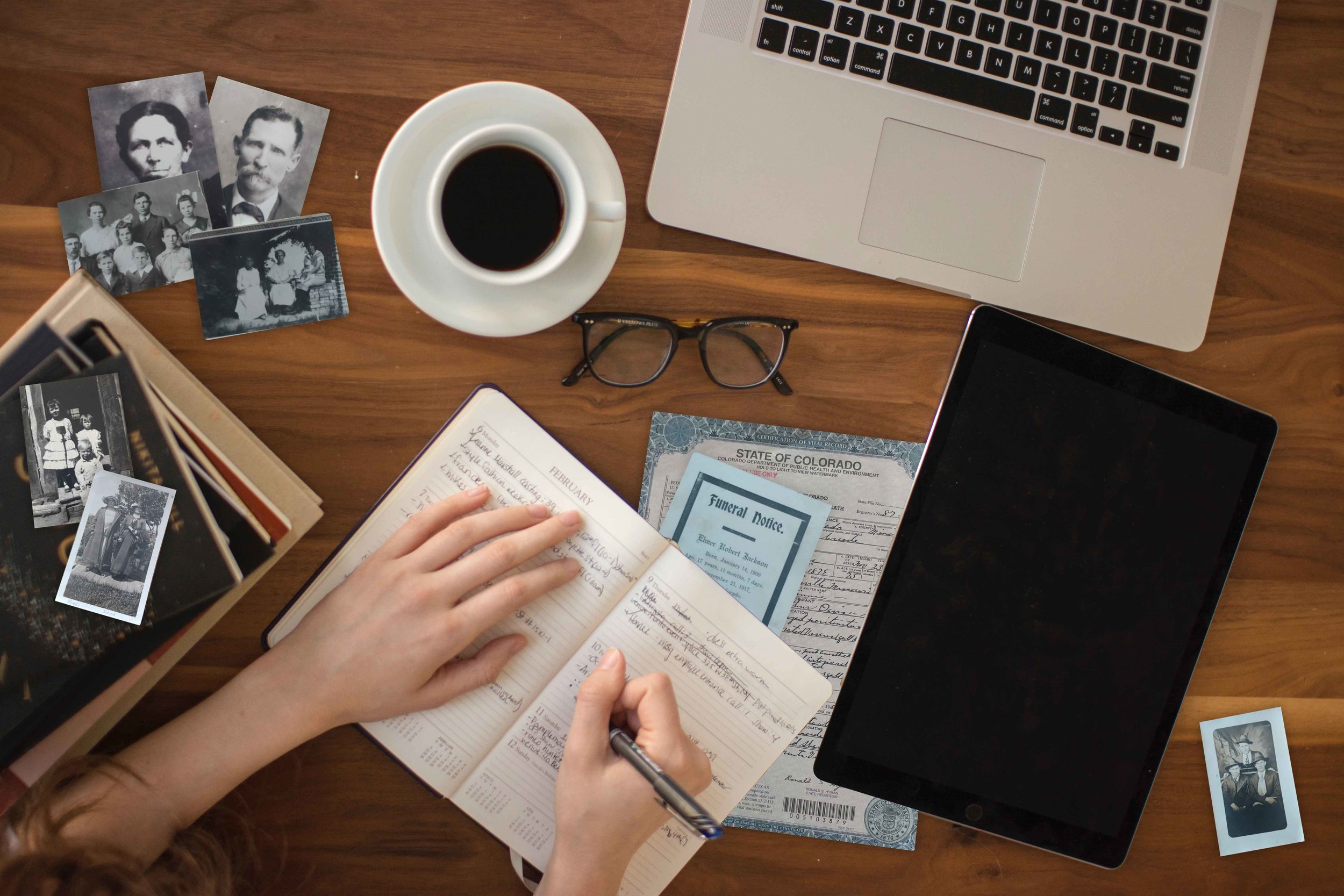 A researcher's desk scattered with old family photographs, an open genealogy notebook mid-entry, a Colorado vital record certificate, reading glasses, and a cup of coffee.