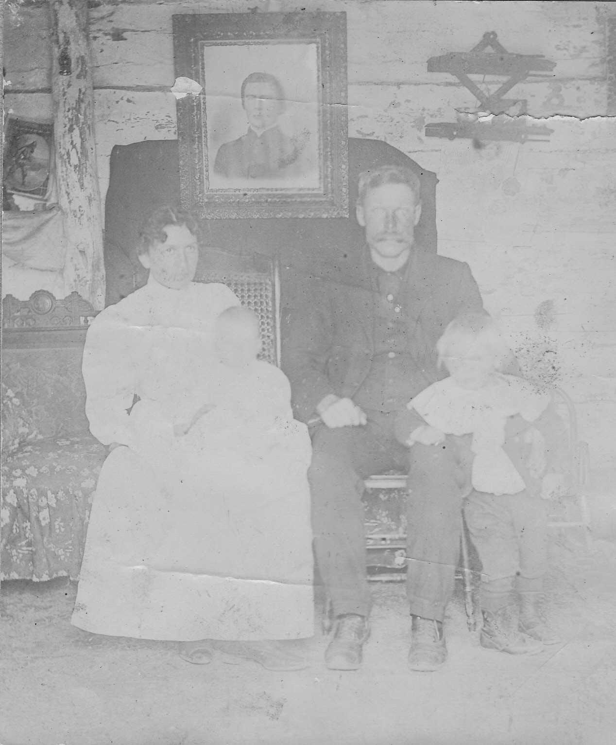 Black-and-white photograph of a man and woman seated inside a rough log cabin, the woman holding an infant in her lap and a young child standing beside the man's knee. A framed Civil War-era portrait of the man hangs on the wall behind them, with tools hanging from pegs on the log wall to the right.