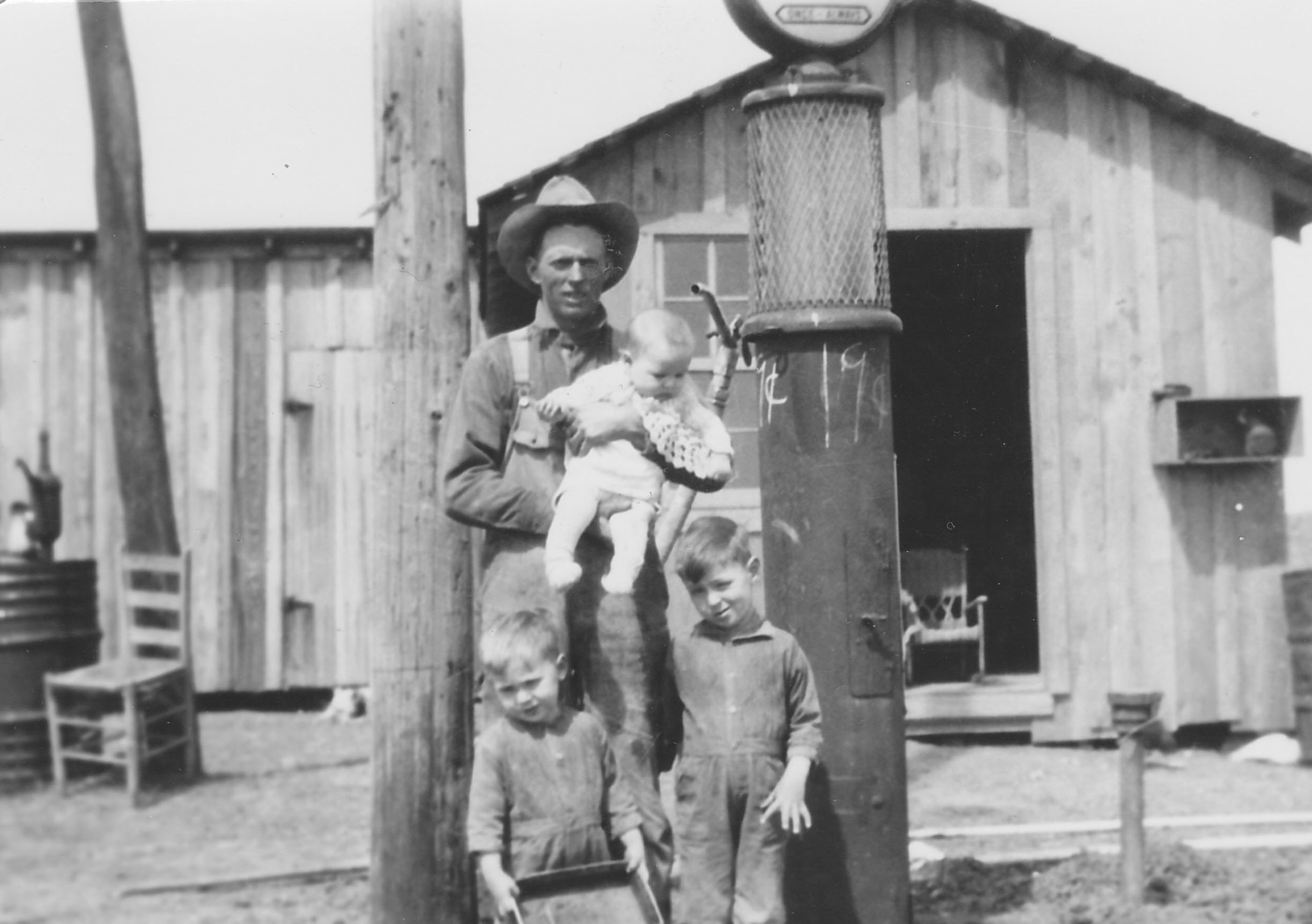 Black-and-white photograph of a man in a wide-brimmed hat and overalls standing beside a gas pump in front of a wood-plank building, holding an infant in a white dress. Two young boys in overalls stand in front of him.