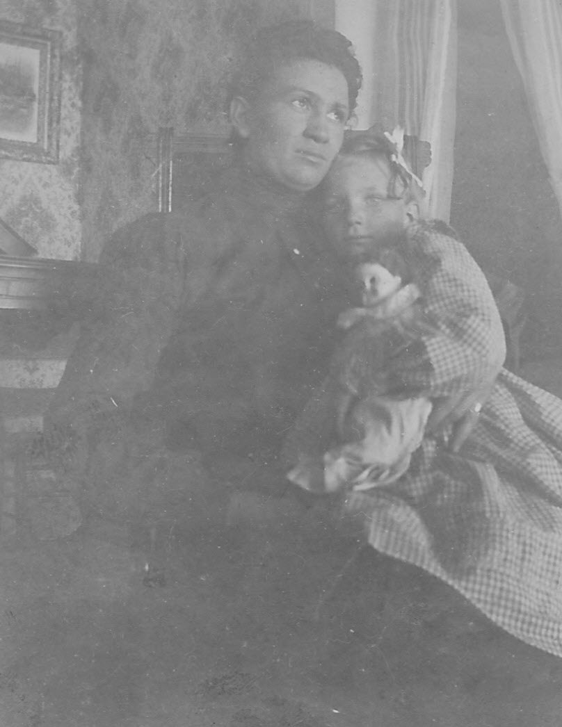 Black-and-white photograph of a young woman seated indoors, holding a small girl close against her, the girl clutching a doll. Patterned wallpaper, a curtained window, and a framed picture are visible behind them.