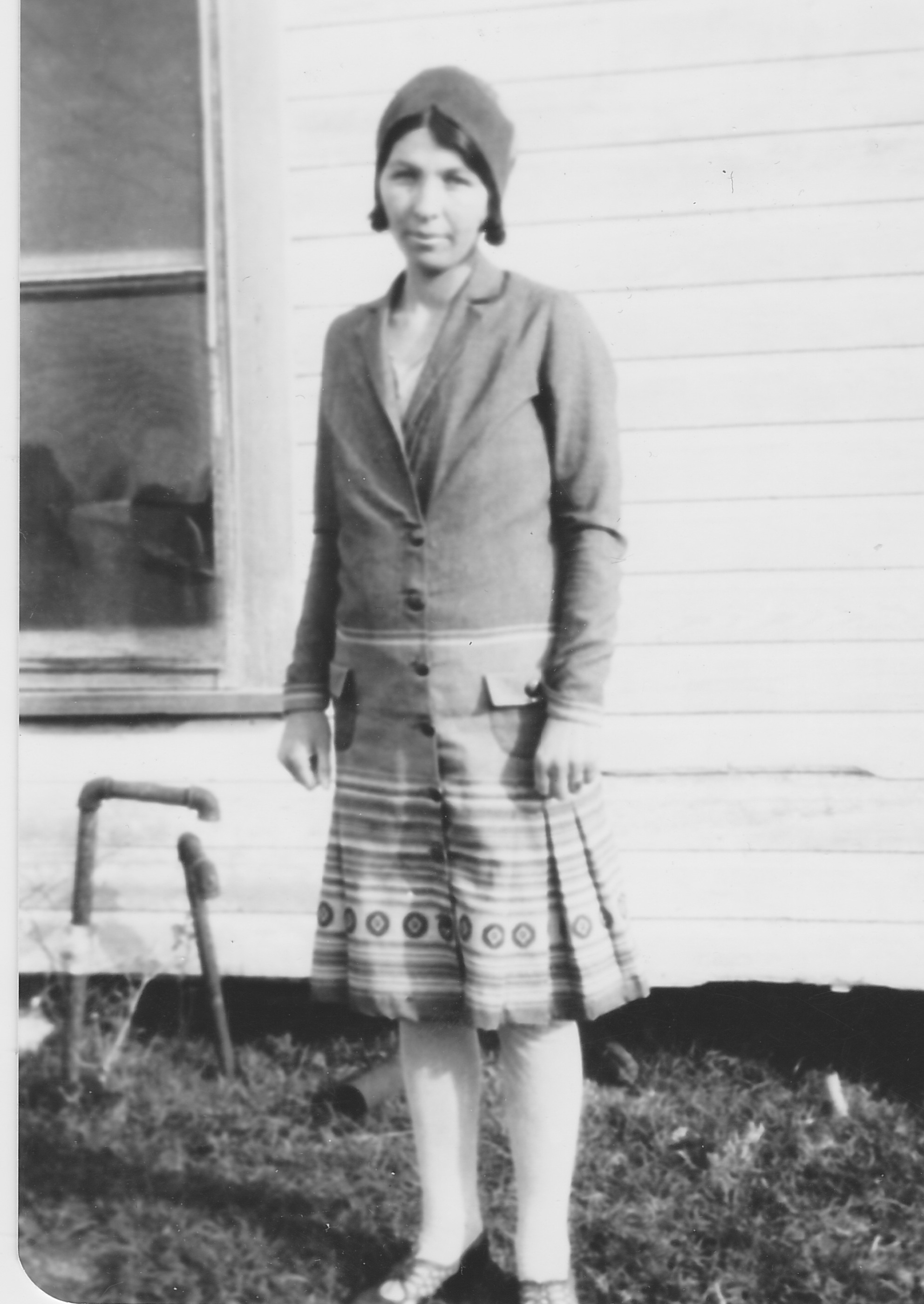 Black-and-white photo of a young Esta Delia Helms standing beside a white clapboard house, wearing a cloche-style hat and a drop-waist coat over a pleated, patterned-hem skirt.