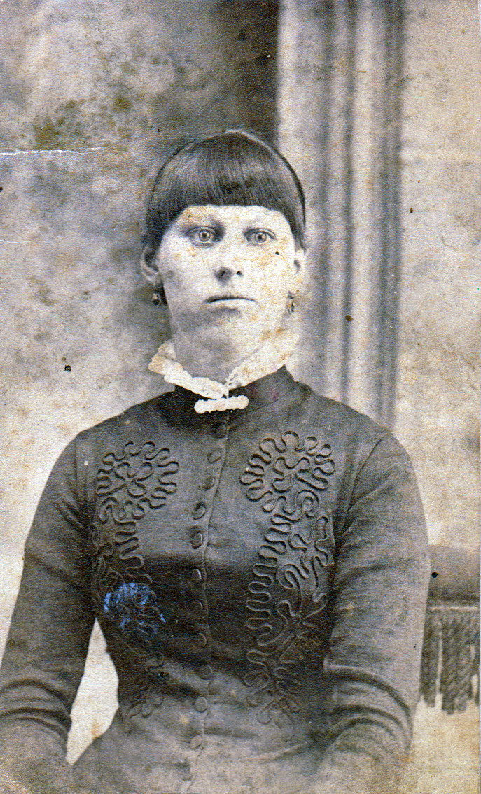 Black-and-white studio portrait of a young Florence Arminda 'Minnie' Staggs, close-cropped, wearing a dark, heavily embroidered dress with a high ruffled white collar and drop earrings. She has blunt-cut bangs and a direct, unsmiling expression.
