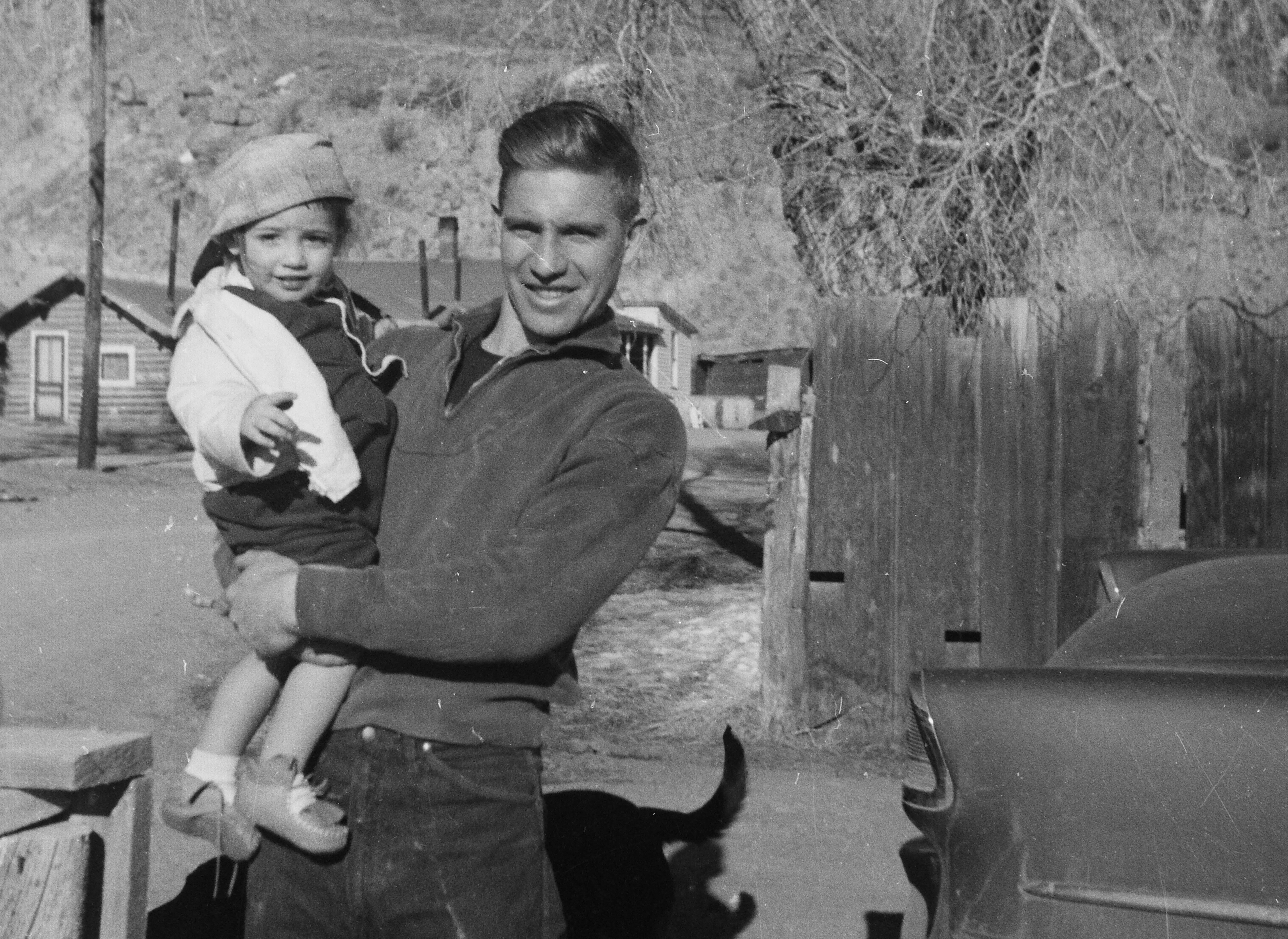 Black-and-white photograph of a young John Richard Jackson holding two-year-old Shannon in Creede, Colorado, circa 1959. Both are smiling; the Creede landscape and buildings are visible in the background.