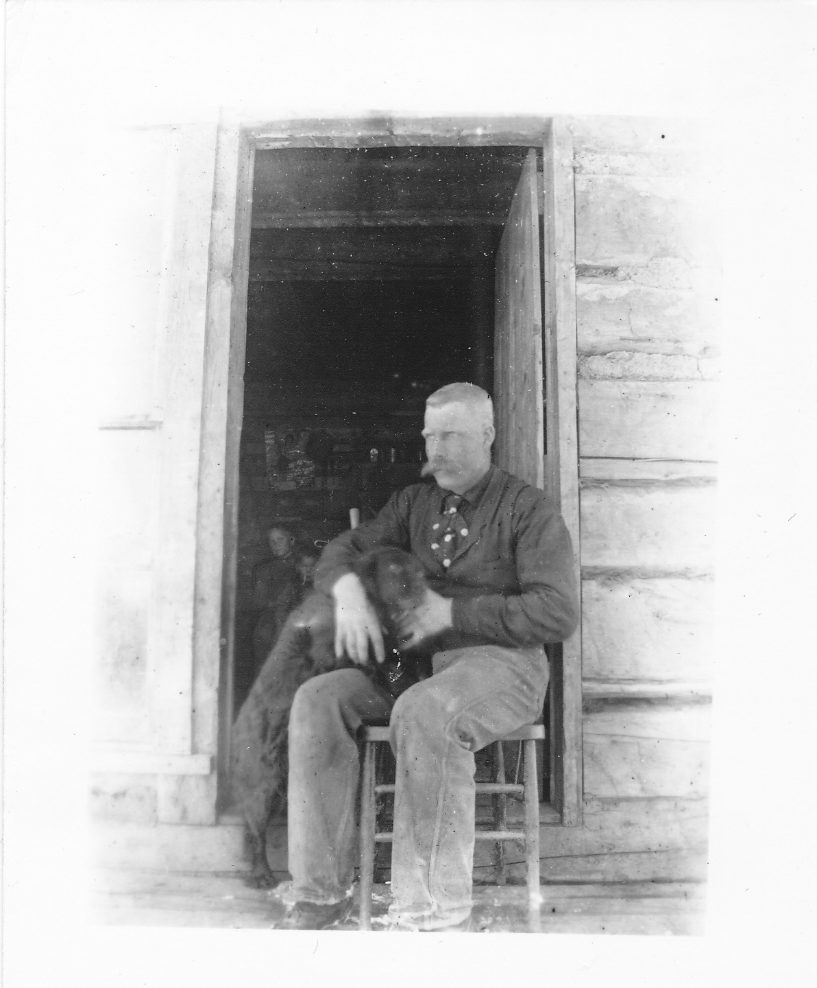 Black-and-white photograph of a man seated on a stool on the front porch, in the open doorway of a log cabin, petting a dog beside him. The exterior log wall with chinking is visible around the doorframe. Two children are visible in the dark interior behind him, through the open doorway.