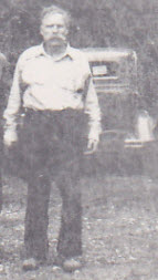 Black-and-white photograph of an older man with a full beard and mustache, standing outdoors in a light-colored button-up shirt and dark trousers. An older-model car or truck is parked behind him.