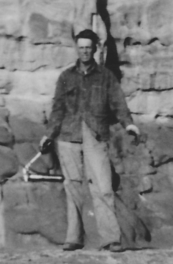 Black-and-white cropped portrait of a young Robert Webster Bates standing in work clothes — a dark button-front shirt and lighter trousers — in front of a tall, layered sandstone rock formation. He holds an unidentified tool in one hand.
