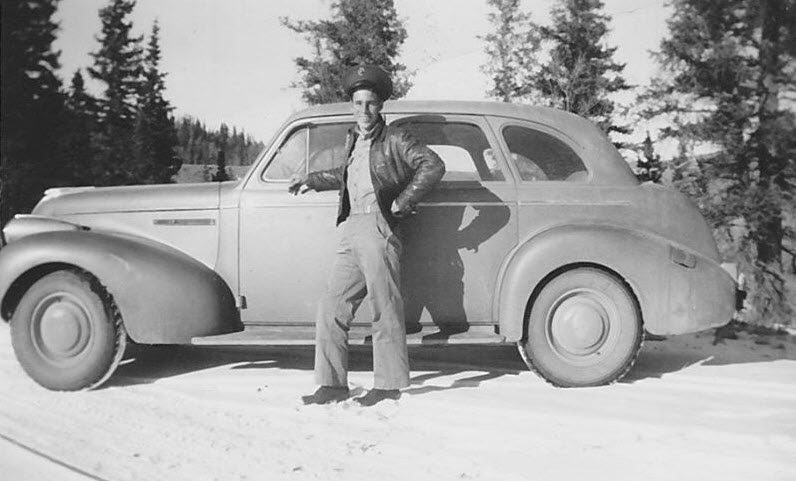 Black-and-white photograph of a young William Charles "Scoot" Jackson standing beside his car in the snow near Creede, one hand braced on the open door, wearing a leather jacket and cap.