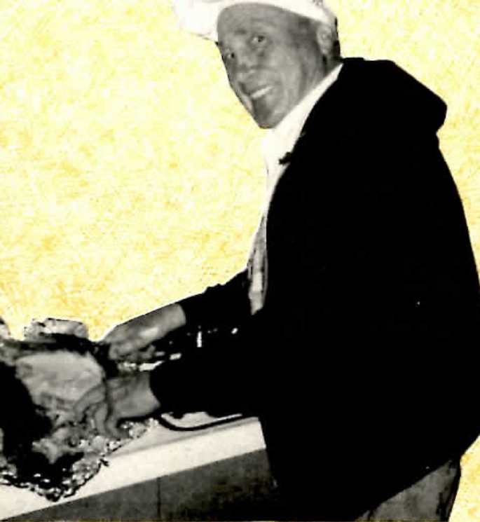 Black-and-white photograph of an older man in a white chef's hat and dark jacket, smiling as he slices a large roast on a kitchen counter.