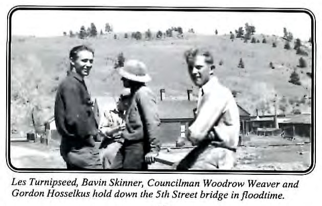 Black-and-white photograph of three men standing together outdoors near a bridge, with a hillside and buildings in the background.