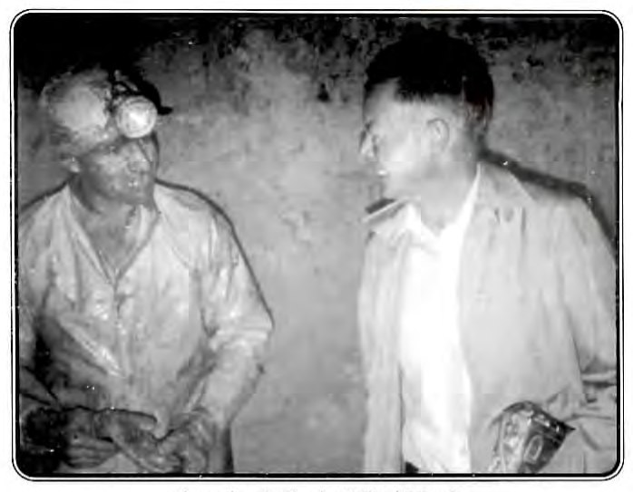 Black-and-white photograph of two men talking underground in a mine, one wearing a miner's cap lamp and work clothes, the other in a light jacket.