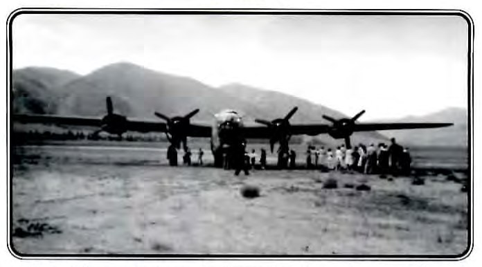 Black-and-white photograph of a four-engine military bomber parked on an open dirt airstrip, with a crowd of onlookers gathered beneath it.