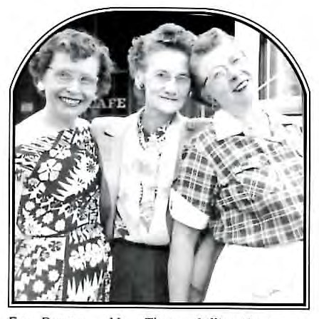 Black-and-white photograph of three women standing close together outdoors, smiling, in front of a café building.