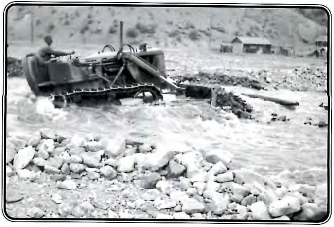 Black-and-white photograph of a man on a bulldozer working in a flooded, rock-strewn creek bed.