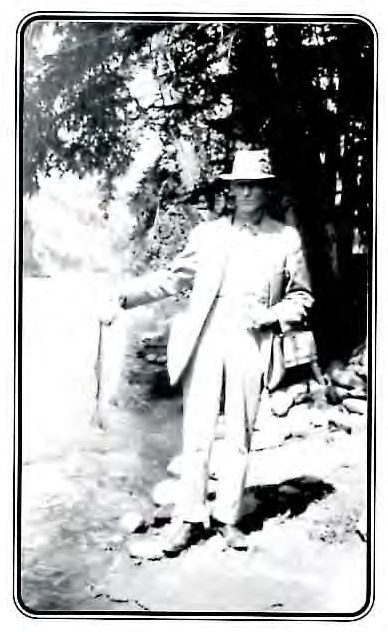 Black-and-white photograph of a man in a light suit and hat standing beside a mountain stream, holding a fishing creel.