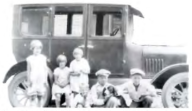 Black-and-white photograph of six children posed in front of a Model T-era automobile, three standing at left, one seated holding a small dog, and two boys seated at right.