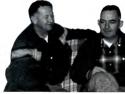 Black-and-white photograph of two men seated close together outdoors, one with his arm around the other's shoulder.