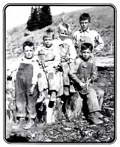 Black-and-white photograph of five boys posed together outdoors on a hillside, some standing and some seated on tree stumps, dressed in overalls and work clothes.
