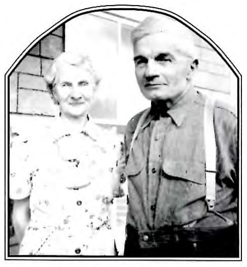 Black-and-white photograph of an older man and woman standing together outdoors in front of a brick building.