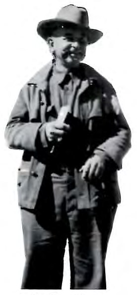 Black-and-white photograph of a man in a wide-brimmed hat and canvas field jacket, standing outdoors.