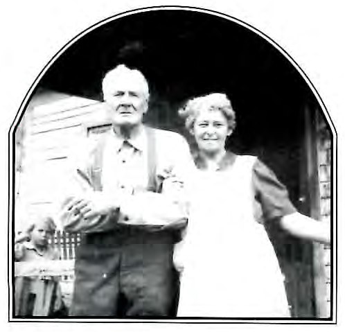 Black-and-white photograph of a man and woman standing arm in arm outdoors in front of a wood building, with a child partly visible at their side.