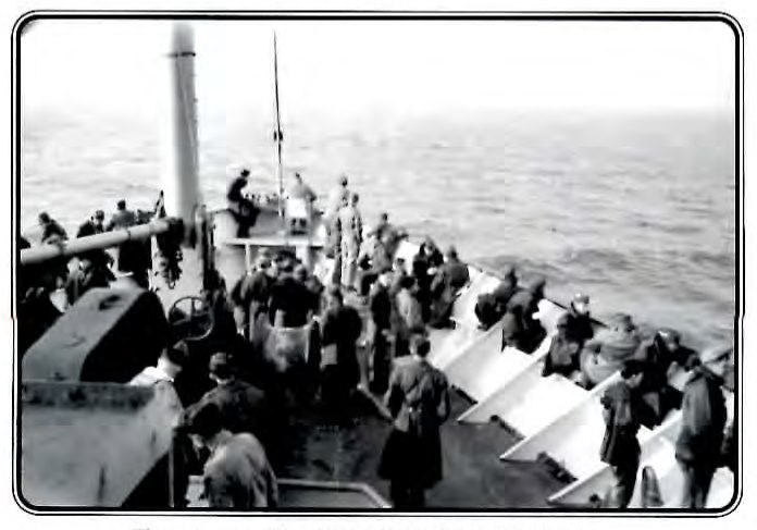 Black-and-white photograph taken from the deck of a troop transport ship, looking down at soldiers filing along a gangway toward the rail.