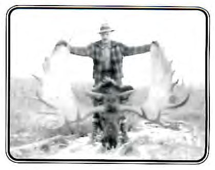 Black-and-white photograph of a man standing behind a large moose head mounted on a post, holding its antlers, in a snowy outdoor setting.