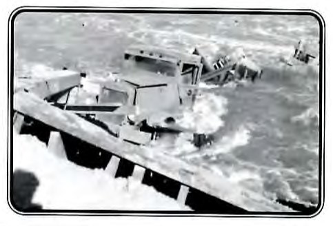 Black-and-white photograph of a wrecked truck cab lying in floodwater beside a collapsed bridge.
