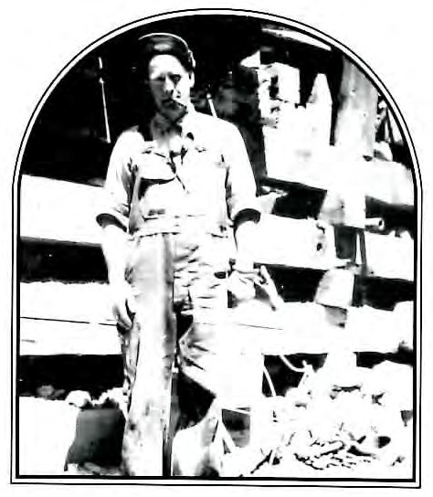 Black-and-white photograph of a man in coveralls standing in front of stacked lumber.