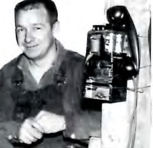 Black-and-white photograph of a man seated beside a wall-mounted crank telephone.