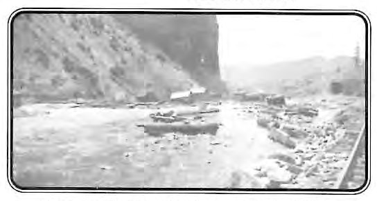 Black-and-white photograph of a flooded creek gorge through Stringtown above Creede, Colorado, with debris and rubble along the banks and a damaged building visible in the distance.