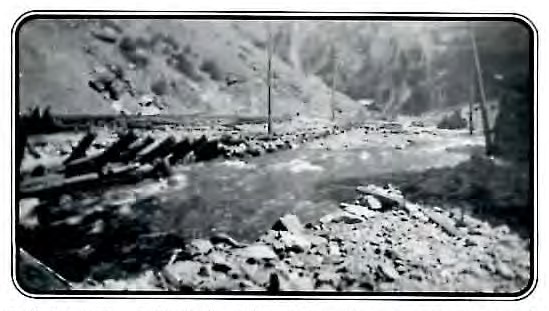 Black-and-white photograph of a rock- and debris-strewn creek bed running through a steep mountain gorge, with scattered timber wreckage along the banks.