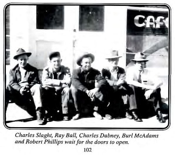 Black-and-white photograph of five young men seated together on a sidewalk in front of a café building.