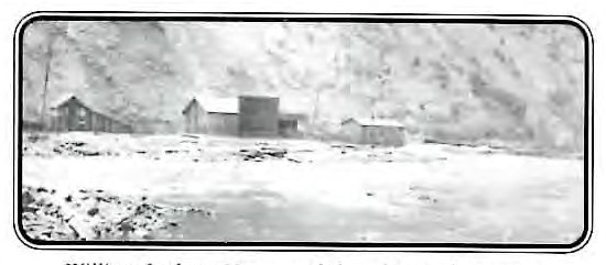 Black-and-white photograph of three wood-frame buildings standing in a snow-covered creek bottom below steep mountainsides, with floodwater visible in the foreground.
