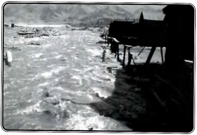 Black-and-white photograph of a swollen creek rushing past a row of buildings on stilts.