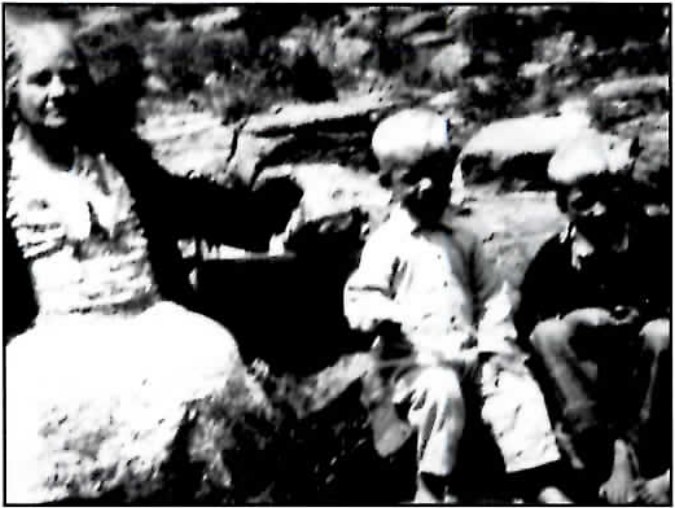 Black-and-white photograph of a heavyset older woman seated outdoors on a rock ledge with two children beside her.