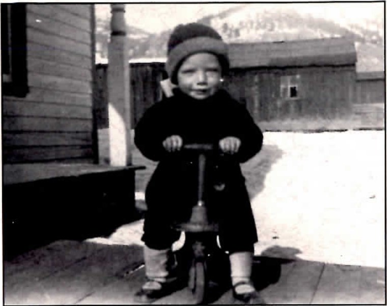 Black-and-white photograph of a small child in a coat and knit cap riding a toy pedal car on a wooden porch, in front of a plank building.