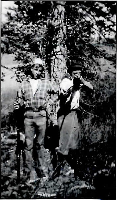 Black-and-white photograph of two teenage boys in hunting gear at a campsite, one aiming a rifle and the other bent over a burro laden with camp gear.