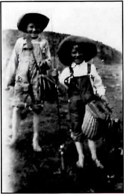 Black-and-white photograph of two barefoot boys in overalls, one holding a stringer of fish and the other a woven fishing creel.