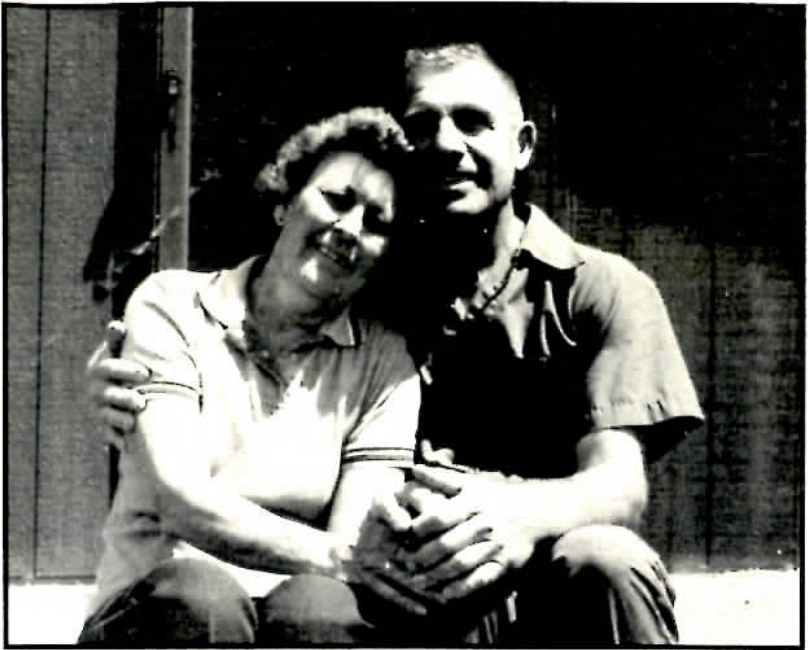 Black-and-white photograph of a man and woman embracing, seated close together and smiling at the camera.