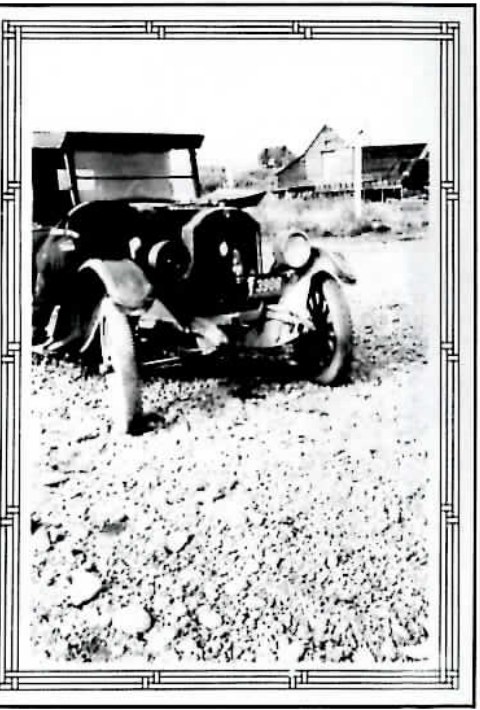 Black-and-white photograph of an early automobile parked in a dirt yard, framed in a decorative border, with farm buildings behind it.