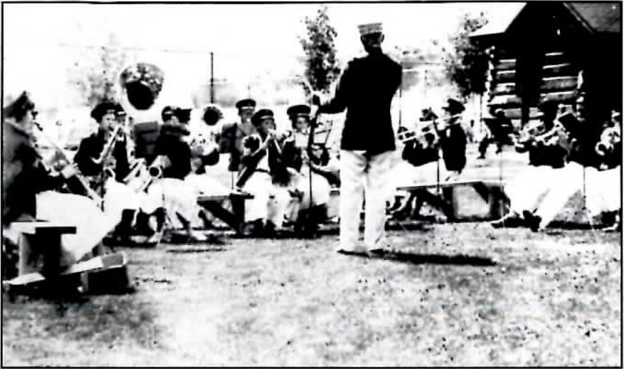 Black-and-white photograph of a children's marching band posed outdoors in uniform, a conductor at the front with his baton raised.