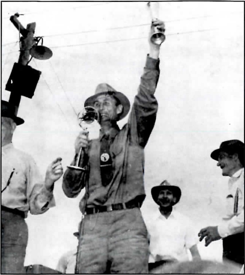 Black-and-white photograph of a man in a hat holding a trophy cup aloft with one hand and drinking from a glass with the other, flanked by two other men, one watching and one laughing.
