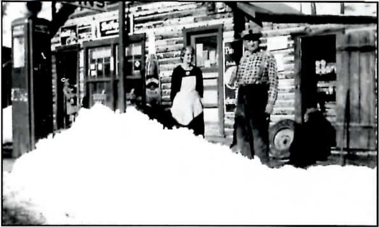 Black-and-white photograph of a small wood-frame service station and store with a gas pump out front, deep snow banked along the walk, and two figures standing near the door.