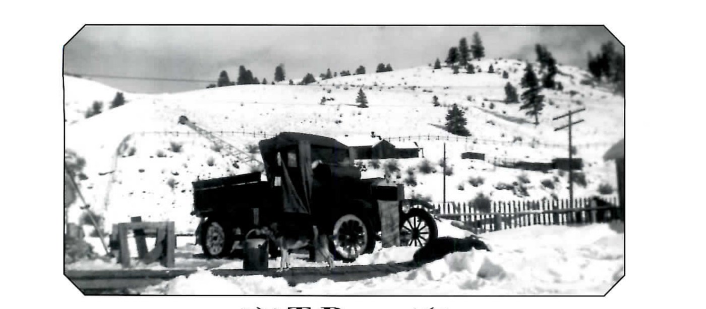 Black and white photograph of an old flatbed truck parked in snow in front of a hillside, with a dragline boom visible behind it