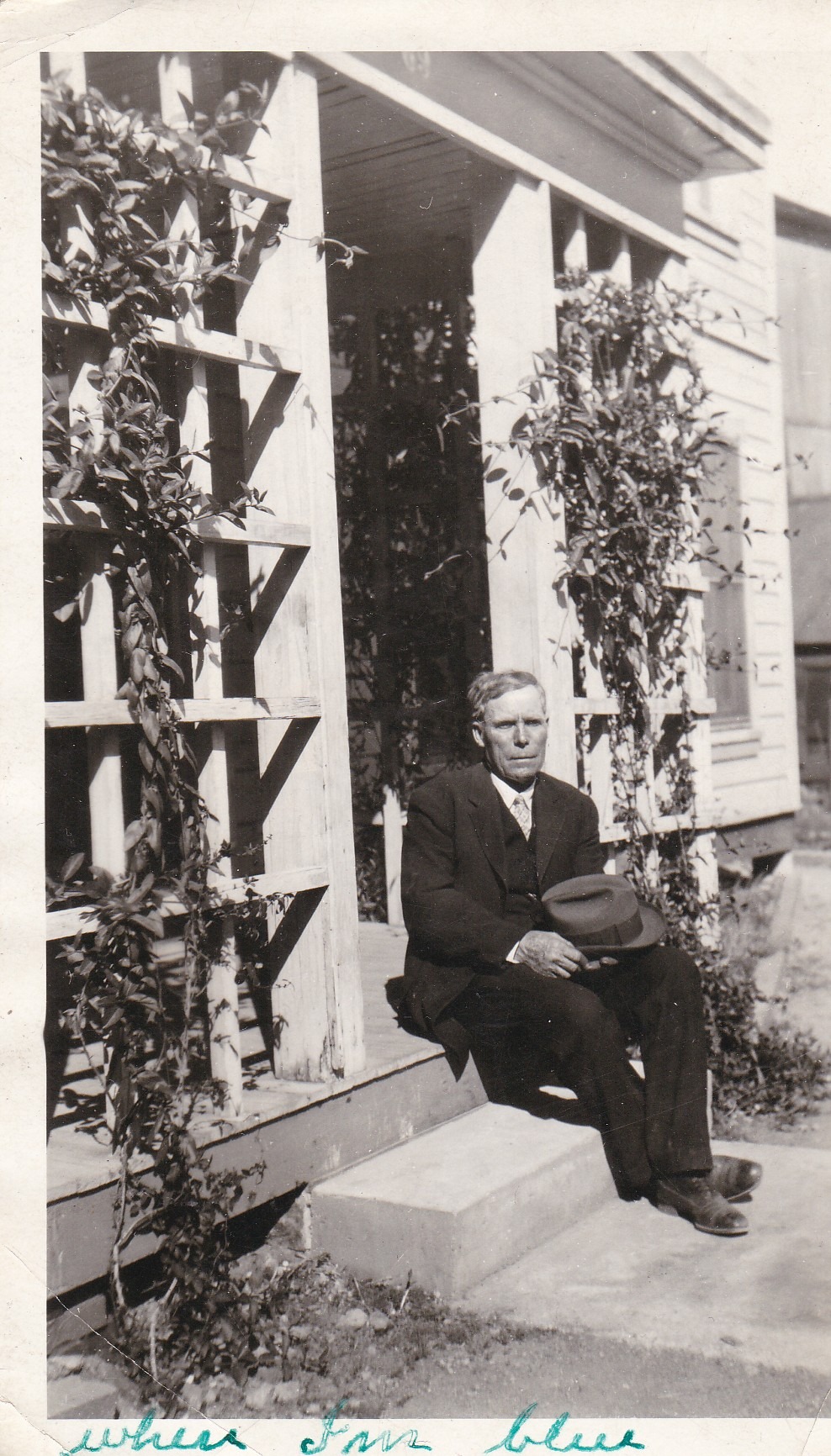 Black-and-white photograph of an older man in a dark suit sitting on a porch step beside trellised vines, holding a hat in his lap, looking directly at the camera.