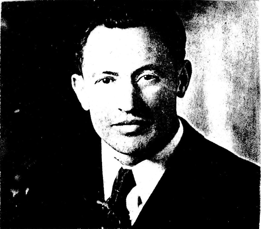 Black-and-white head-and-shoulders portrait of a man with short dark hair in a dark suit, white shirt and dark tie, facing the camera.