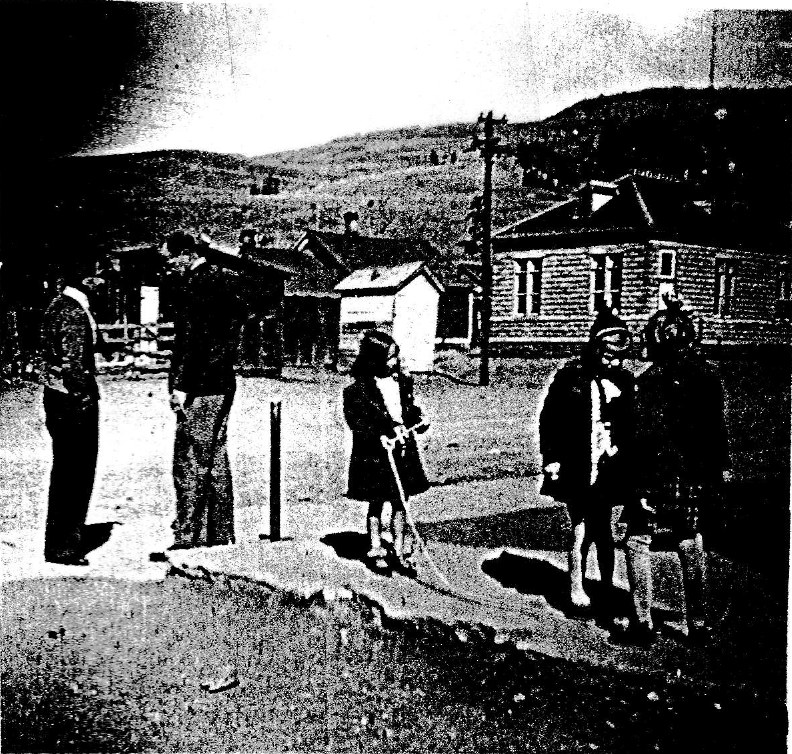 Black-and-white photograph of a dirt street in Creede with two men standing at the left and three small children in coats and knit caps at the right, one girl holding a long rope or handle; a wood-sided building with tall windows and a small white outbuilding stand behind them, with hills beyond.
