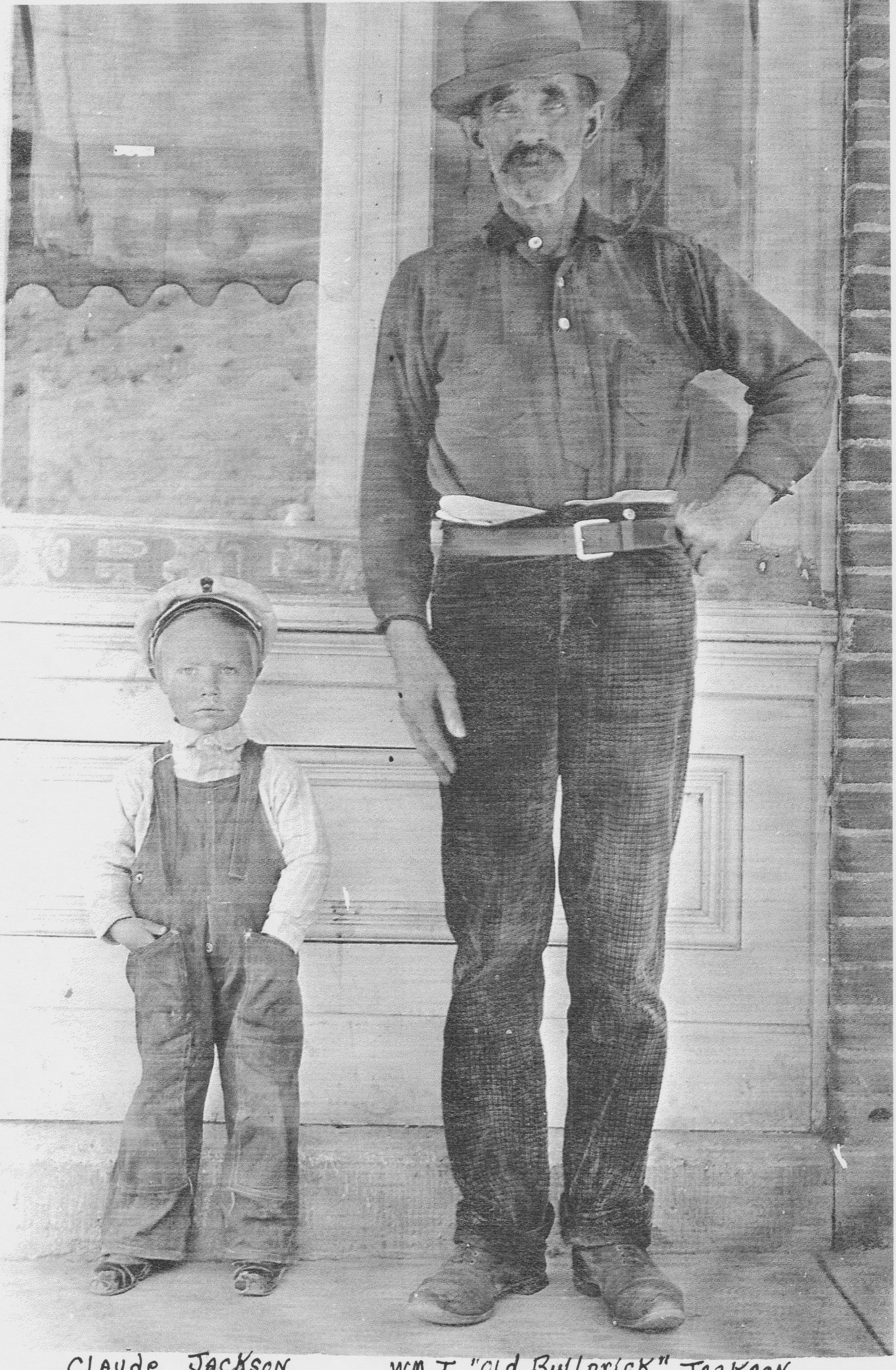 Black-and-white photograph of an older man with a mustache and goatee, wearing a broad-brimmed hat and standing with one hand on his hip, beside a small boy in overalls and a flat cap, in front of a storefront window and brick wall.