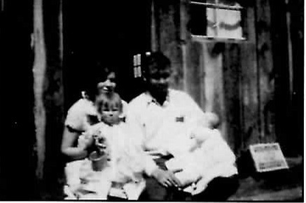 Black-and-white photograph of a woman and a man sitting together in front of a wood cabin doorway, each holding a small child dressed in white.