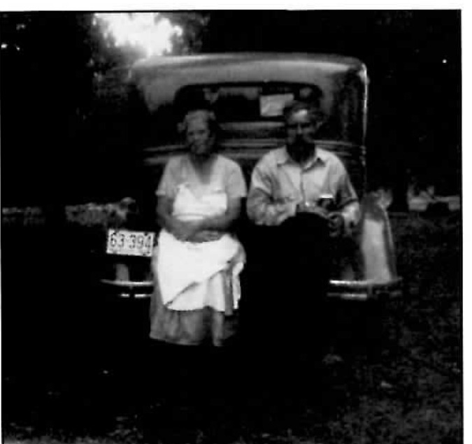 Black-and-white photograph of an older woman in a light dress and apron and an older man in a work shirt, standing together at the rear of a parked automobile at night, lit by a campfire glow to one side.